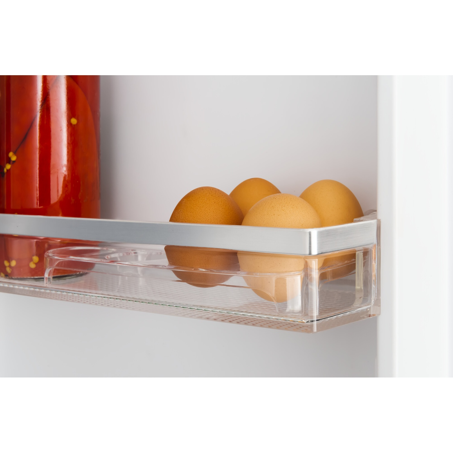 Inside a fridge, four brown eggs sit in a clear plastic container. A jar of red peppers is in the background.