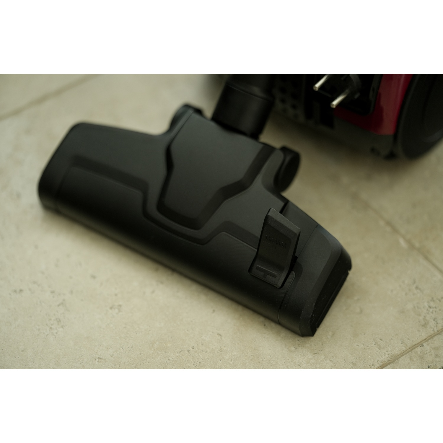 A black vacuum cleaner head on a tiled floor. A red vacuum body and plug are visible in the background.
