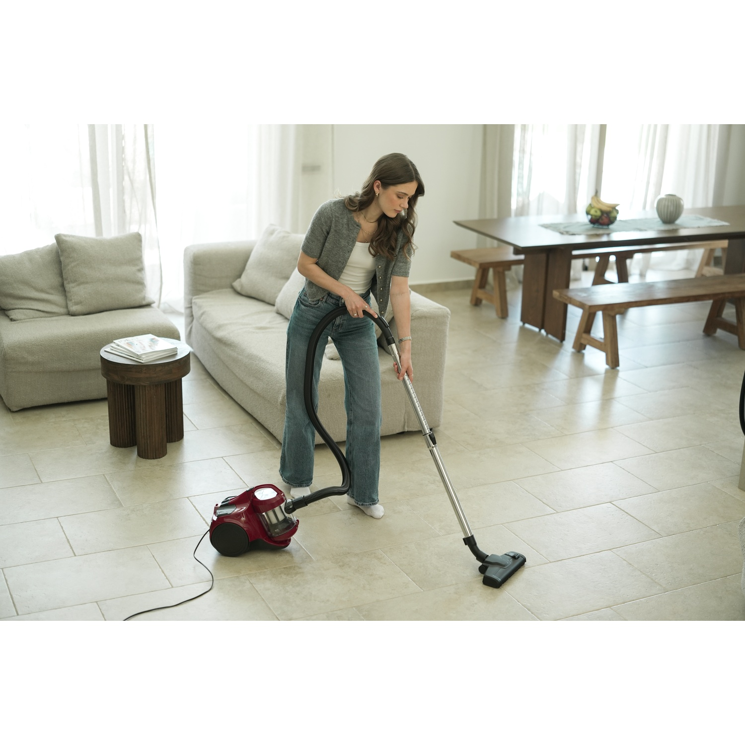 A woman vacuums a tiled floor. She wears jeans and a gray cardigan. A red vacuum cleaner sits nearby.