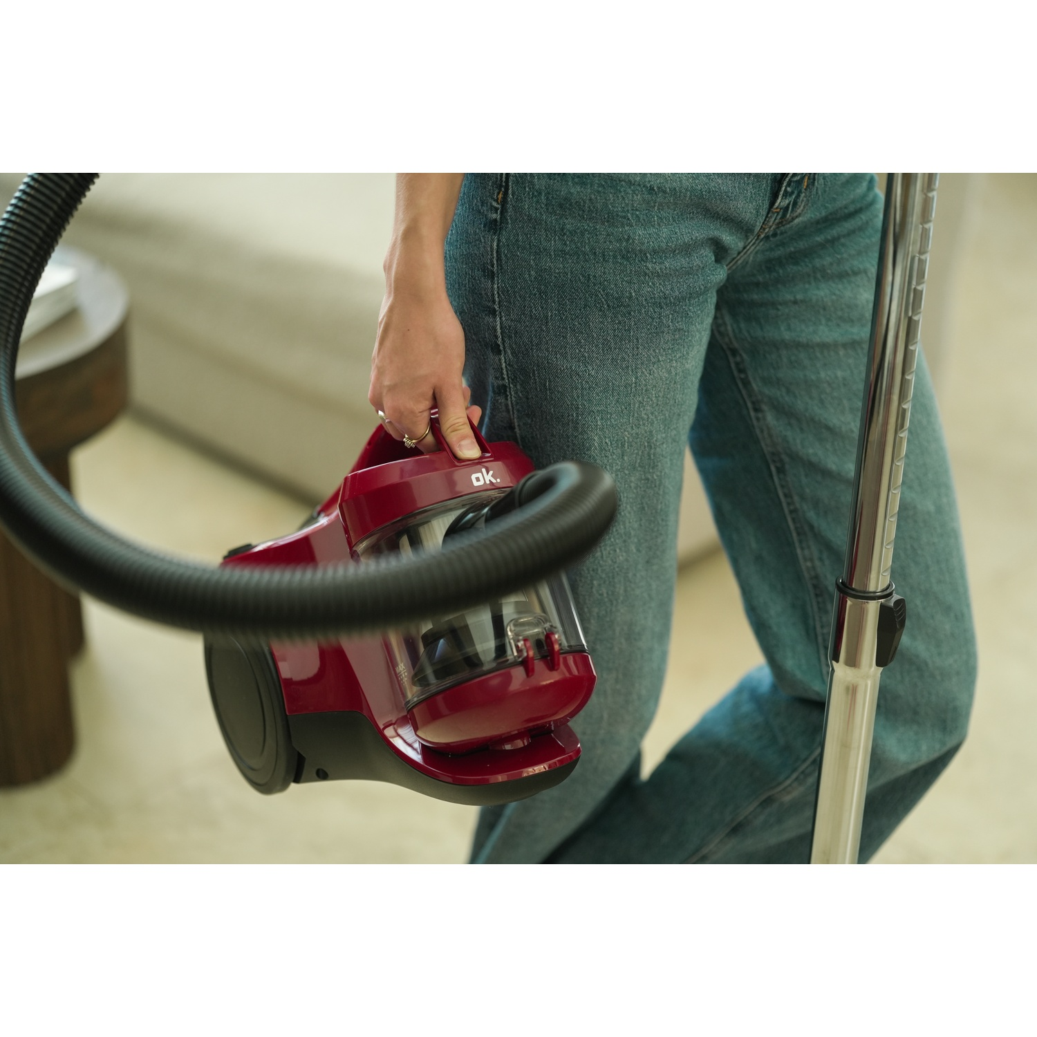 A person holds a red and black vacuum cleaner. Blue jeans and a silver pole are also visible.