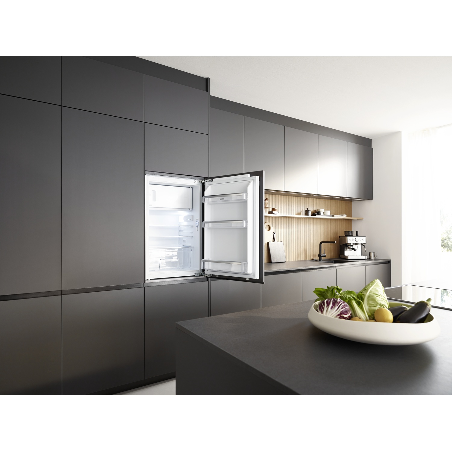A modern kitchen with a fridge open. A bowl of vegetables sits on a countertop. Kitchen cabinets in the background.