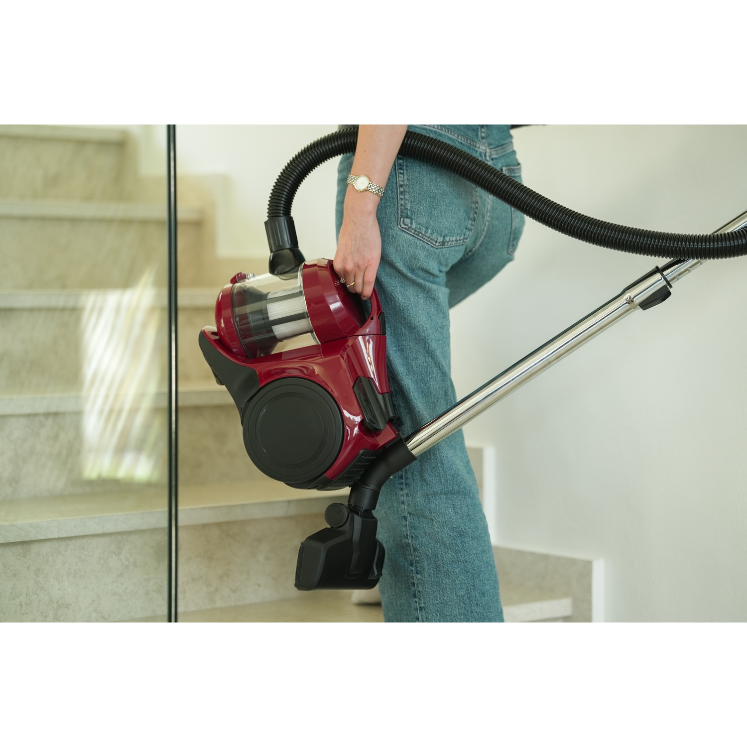 A person in jeans carries a red vacuum cleaner down stairs. The stairs are white with glass paneling.