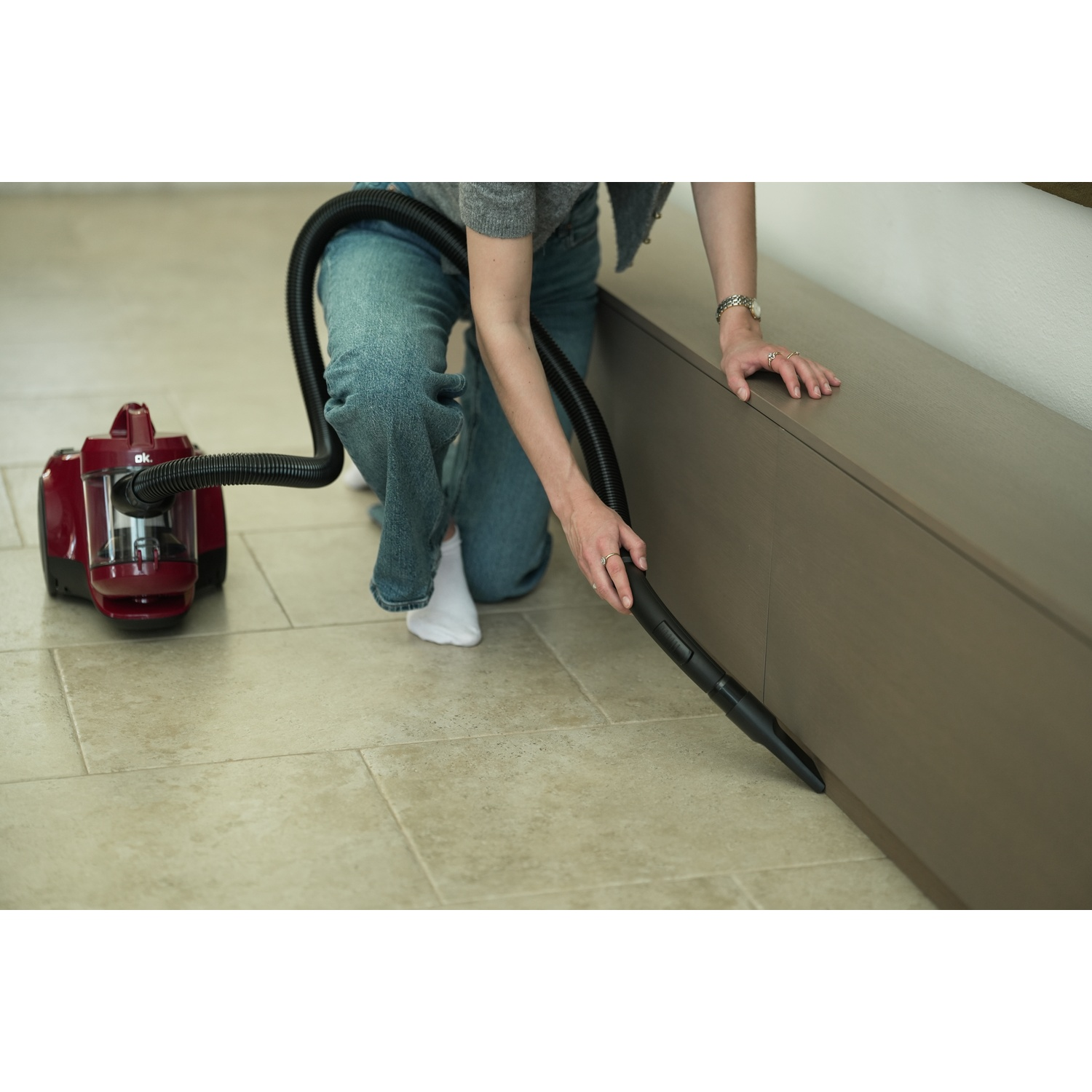 A person vacuums a room. A red vacuum cleaner is on the floor. The person is using the hose to clean near a brown cabinet.