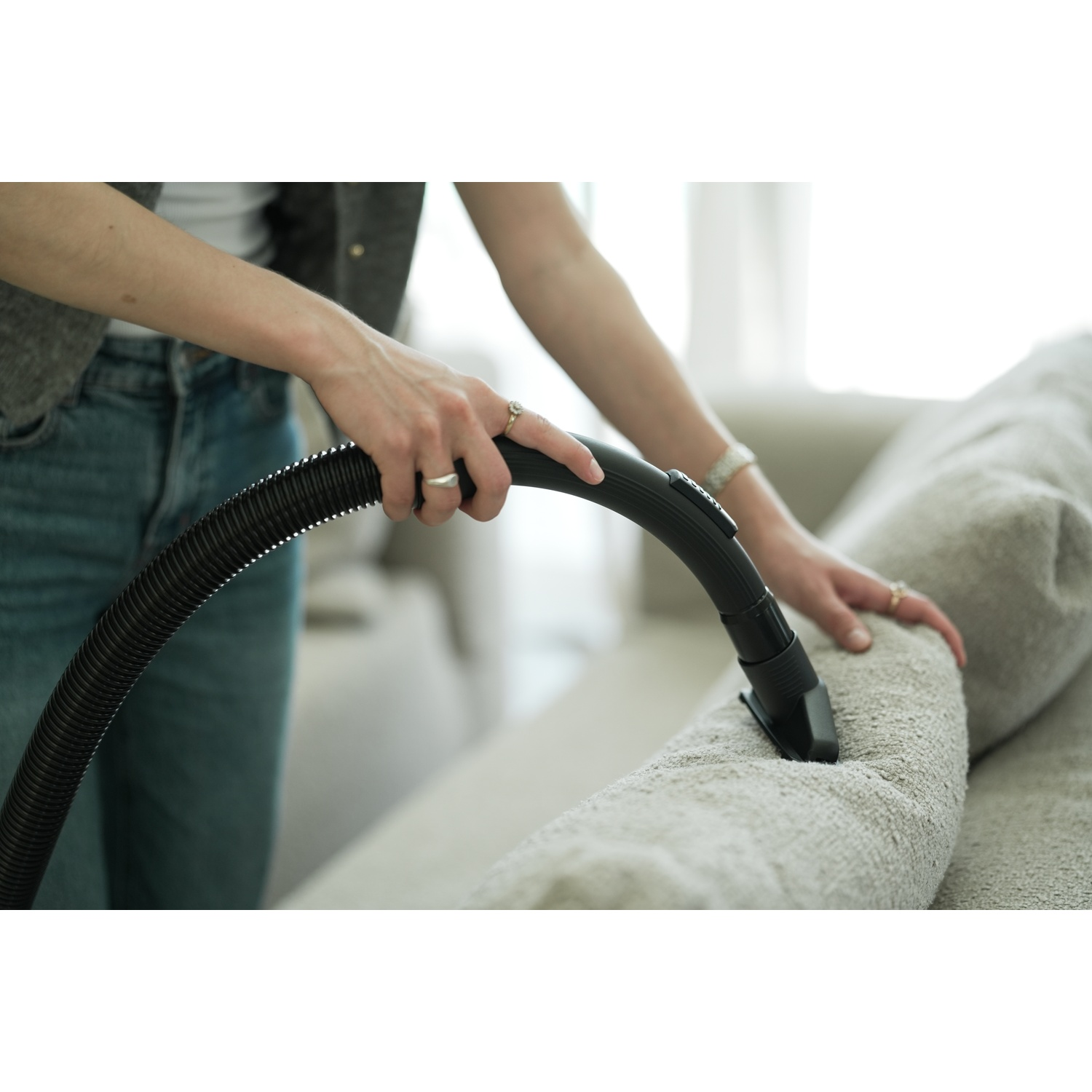 A person vacuums a light-colored couch. The person's hands hold the vacuum hose, which is black. The background is blurred.