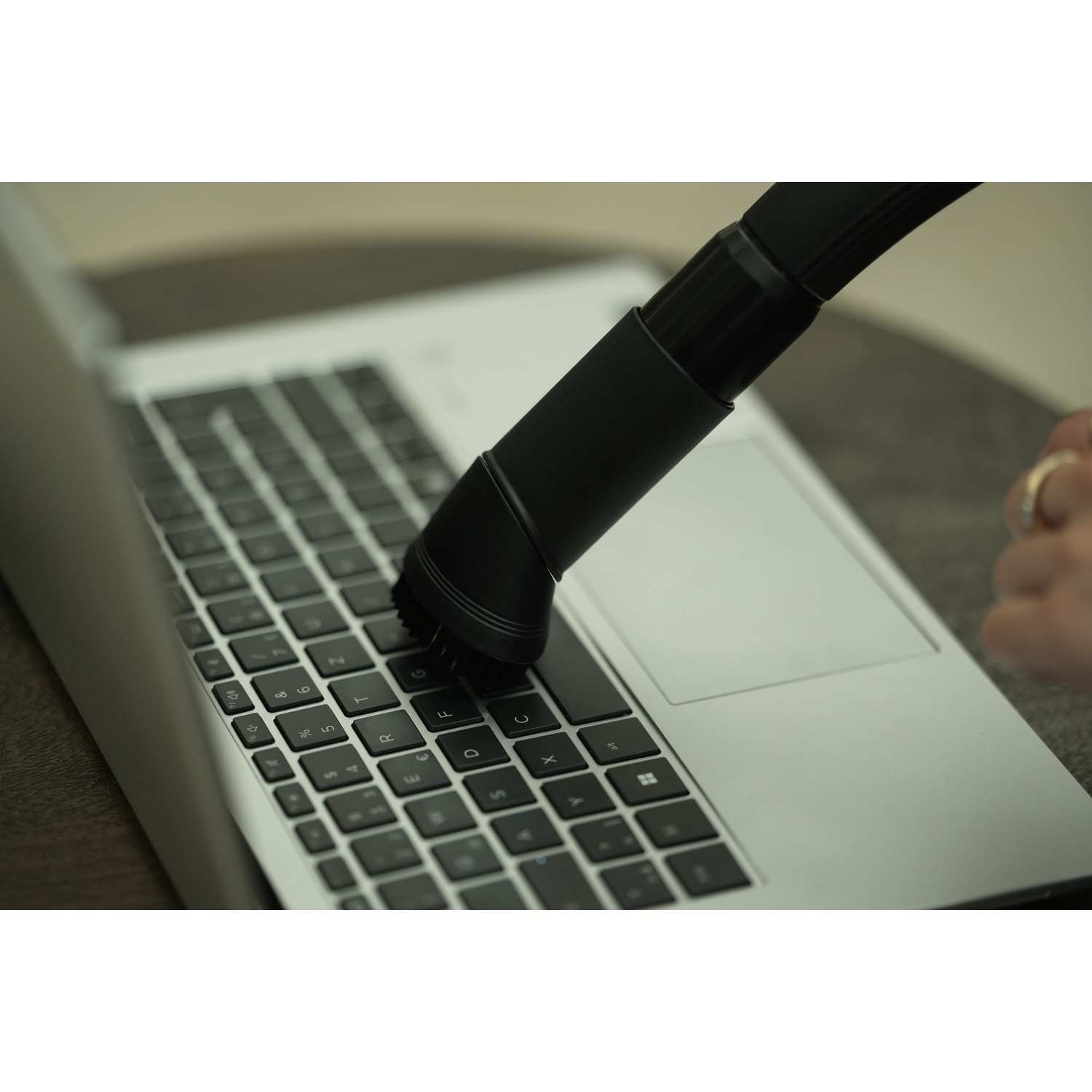 A black vacuum cleaner head cleaning a laptop keyboard. The laptop is silver, and a hand with a ring is visible.