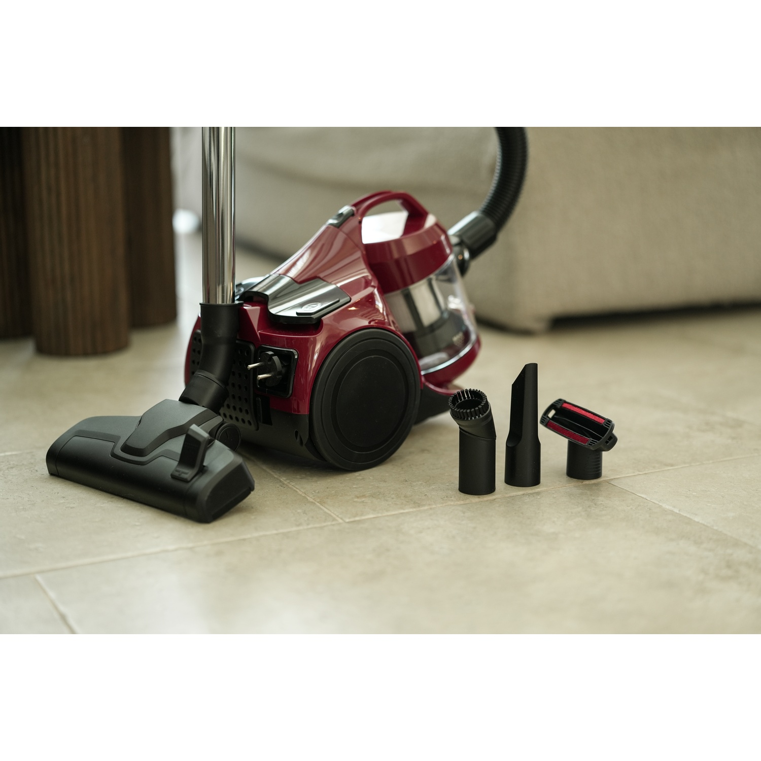 A red vacuum cleaner with black attachments is on a tiled floor. A sofa is in the background.