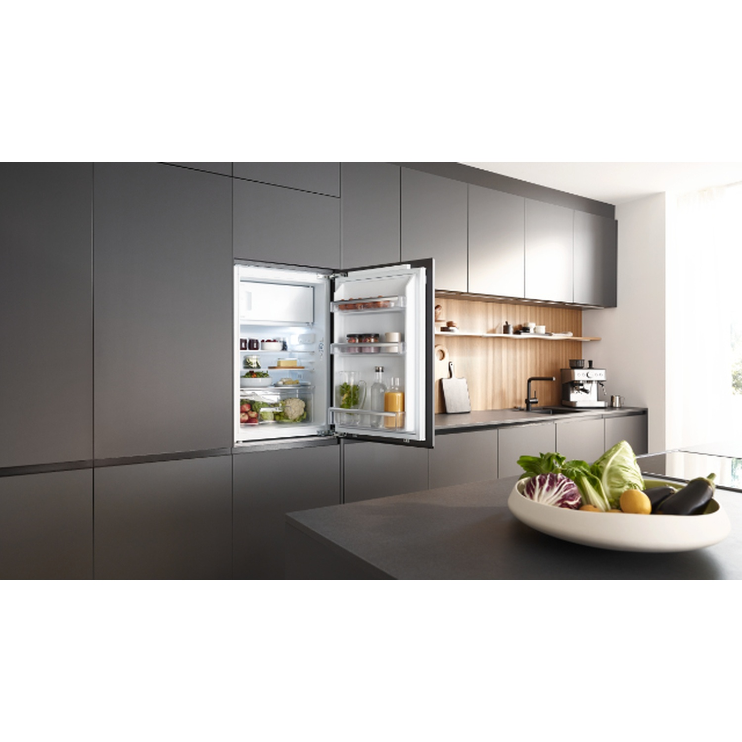 Open fridge in a modern kitchen. Vegetables in a bowl on counter, espresso machine. Grey cabinetry and wooden accents.