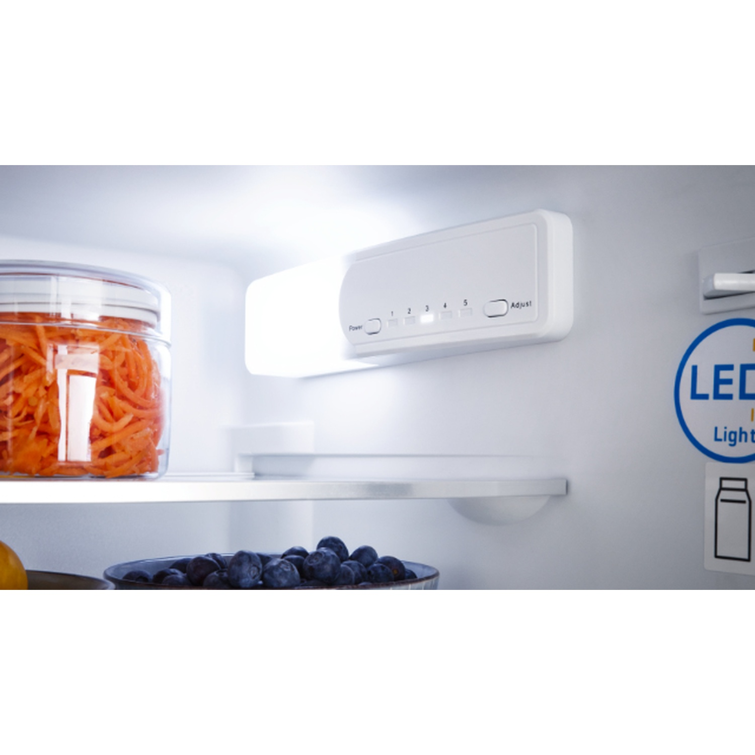 Inside a fridge. A glass jar with orange carrots, blueberries in a bowl, and a light panel with settings are visible.