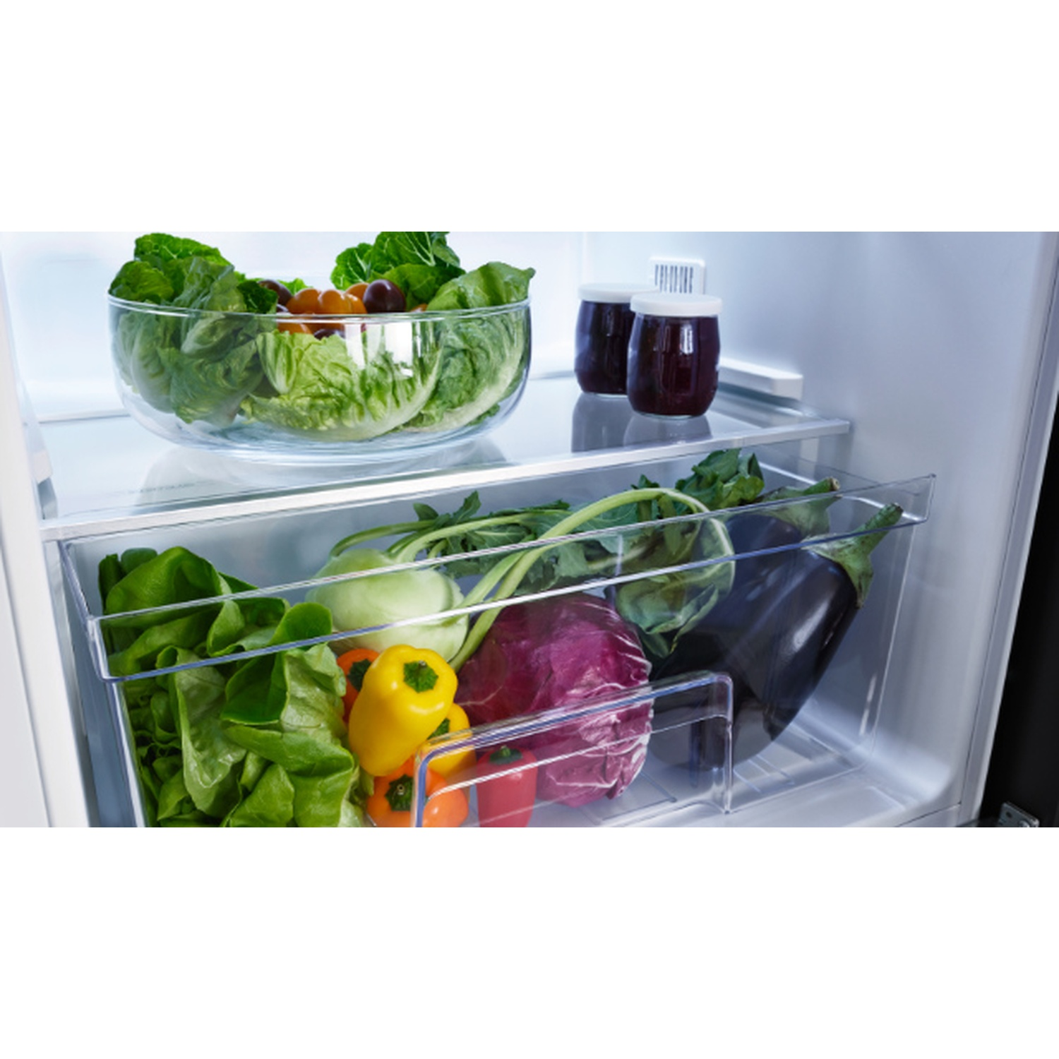 Inside a refrigerator, a glass bowl of salad, jars, and a drawer with vegetables like lettuce, bell peppers, eggplant, and cabbage.