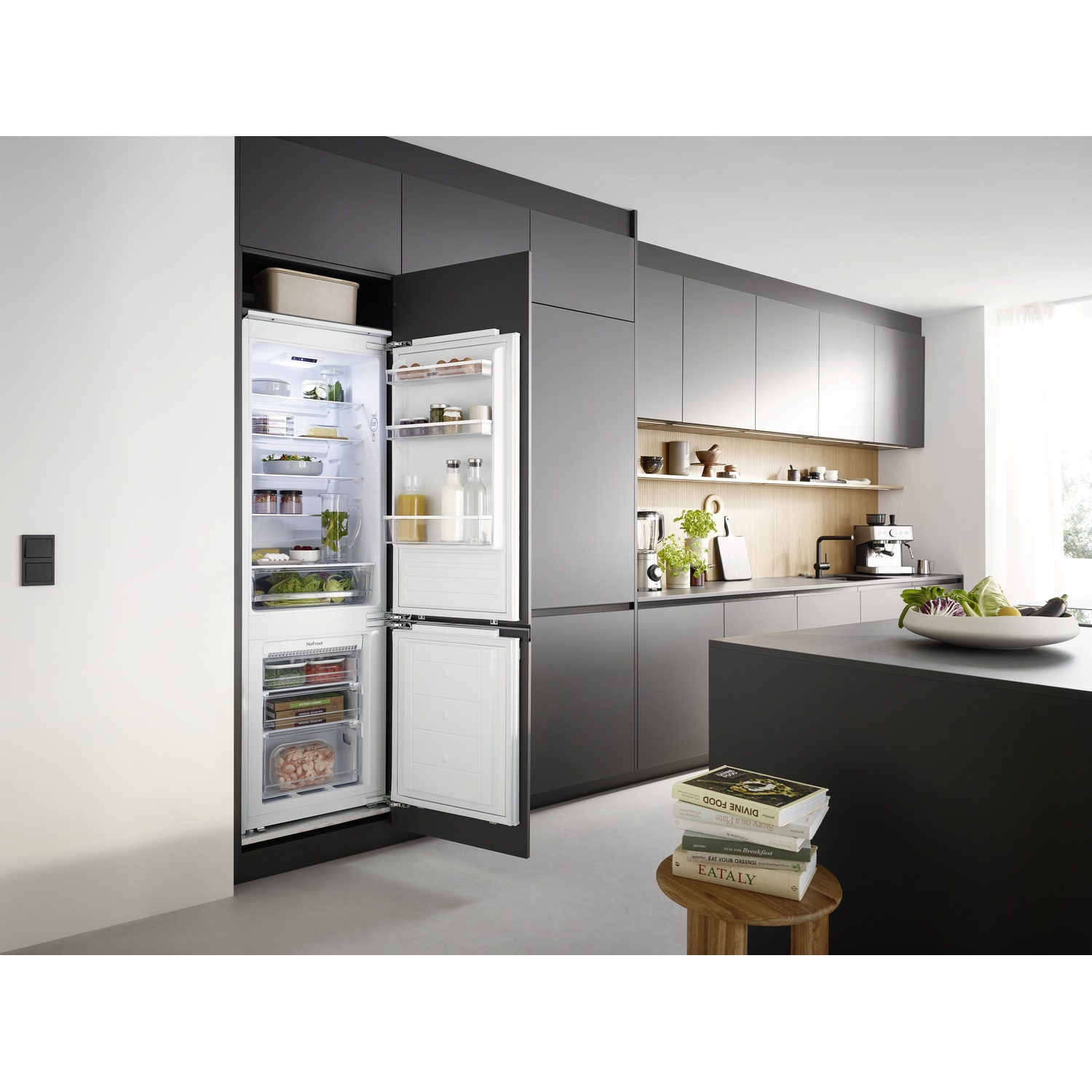 Open fridge shows food. Modern kitchen with island and cookbooks. Black cabinets and white walls.