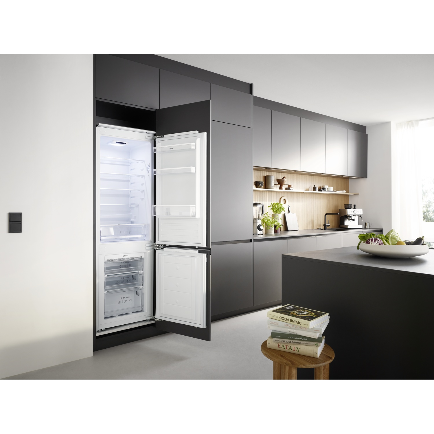 Open fridge in a modern kitchen. Books on a stool, island counter, and cabinets are visible in the background.
