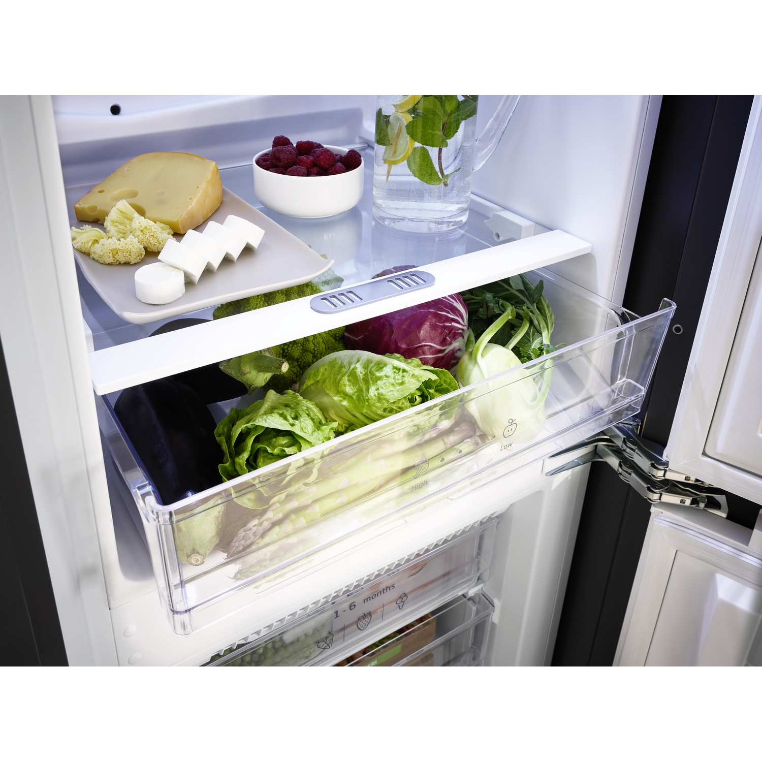Inside a refrigerator: cheese, raspberries, water with lemon and mint, vegetables in a drawer, and eggplants.
