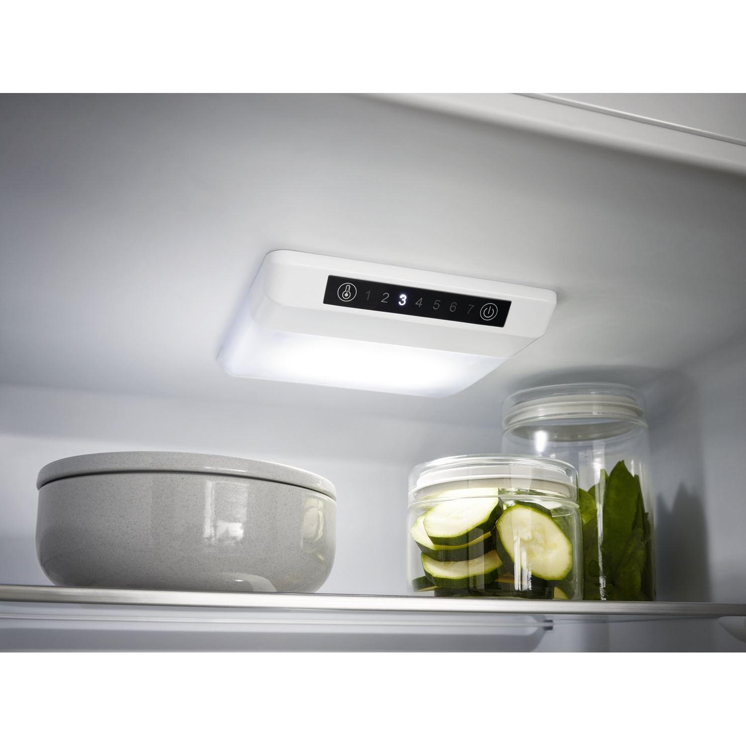Inside a refrigerator: a light panel with numbers, a bowl, two jars with zucchini slices and green leaves.