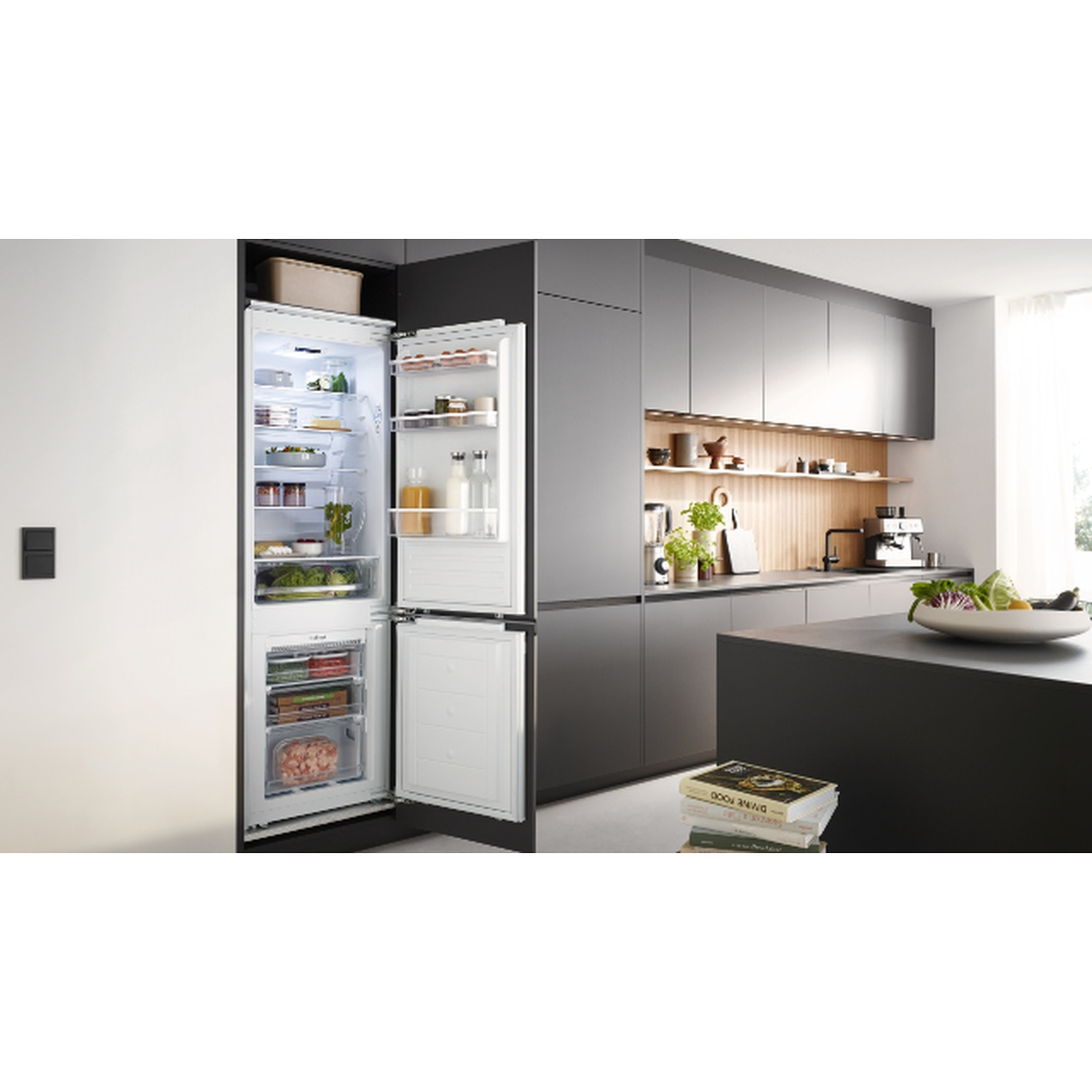 Open fridge in a modern kitchen. Inside, food is visible. Gray cabinets, countertop, and a white wall with light switches are also present.