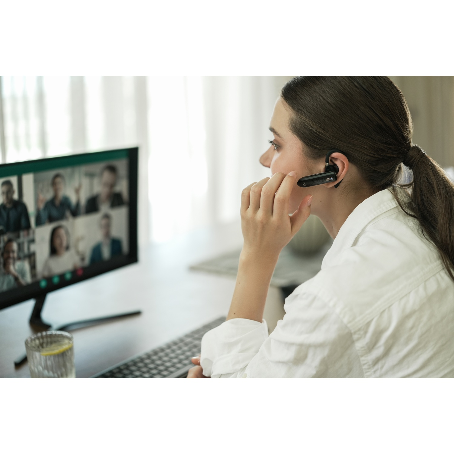 Woman with headset on video call. Monitor shows other people. She wears a white shirt, hand near face.