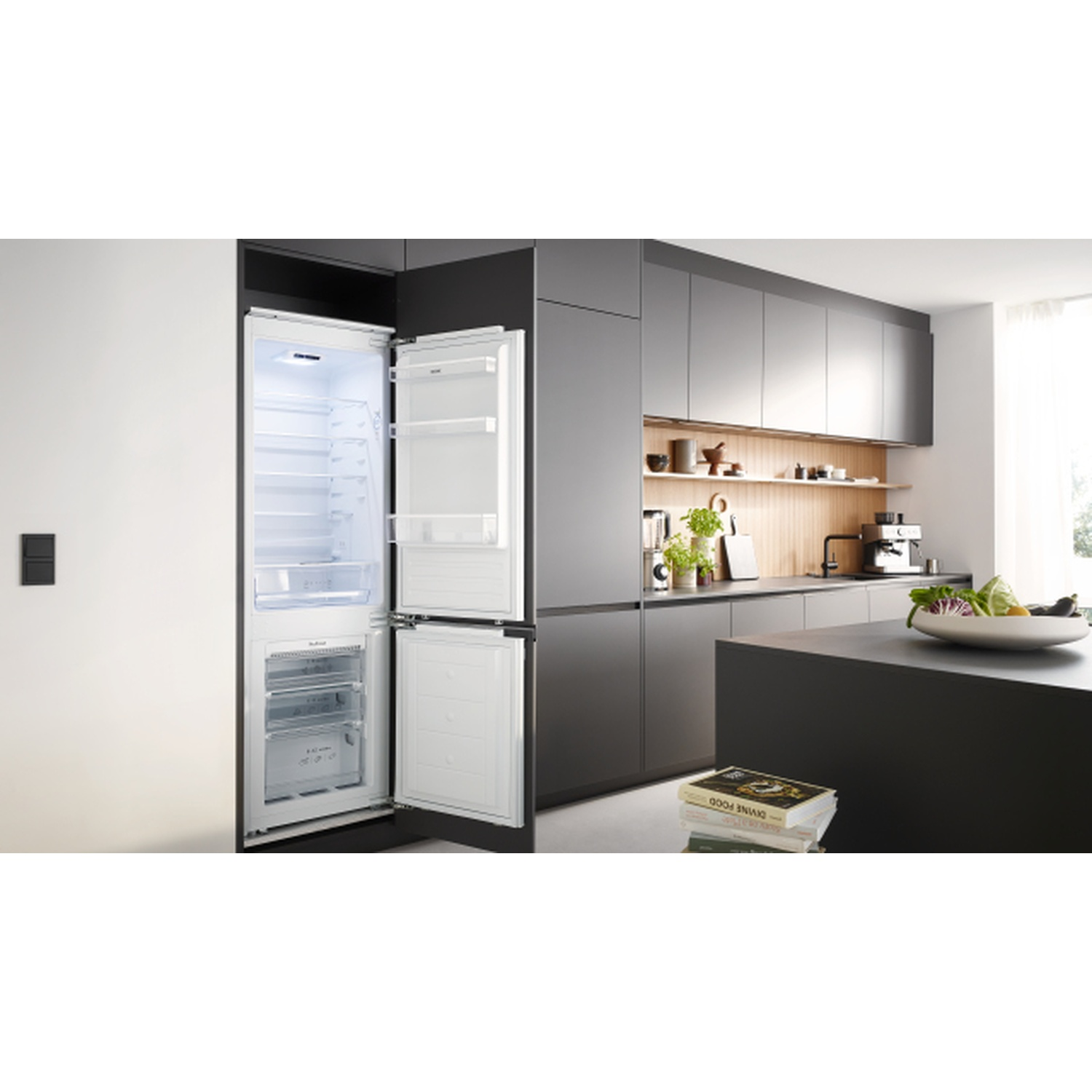 Open white refrigerator in a modern kitchen. Dark gray cabinets, island, and wall. Books on the island, and food.