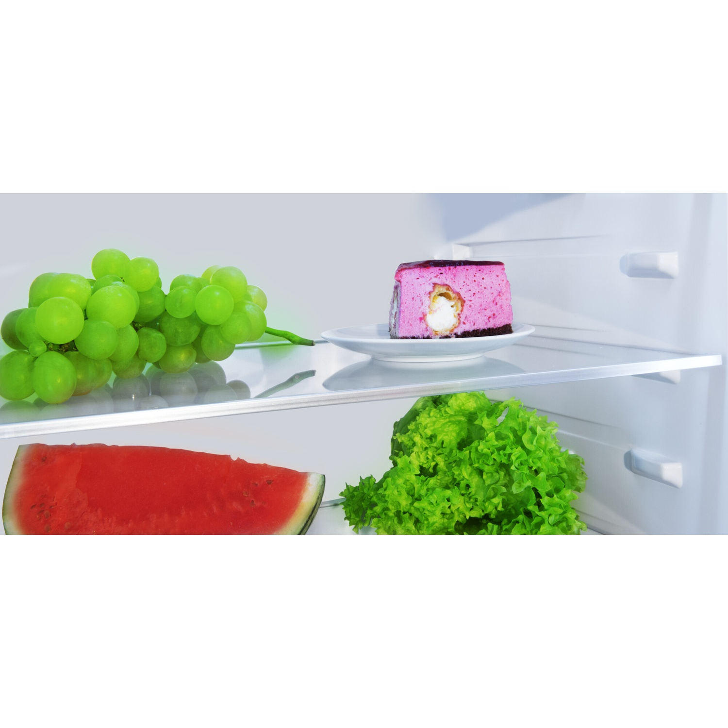 À l'intérieur d'un réfrigérateur : raisins, gâteau, pastèque et laitue. Le gâteau est rose avec une garniture blanche. Le frigo est blanc.