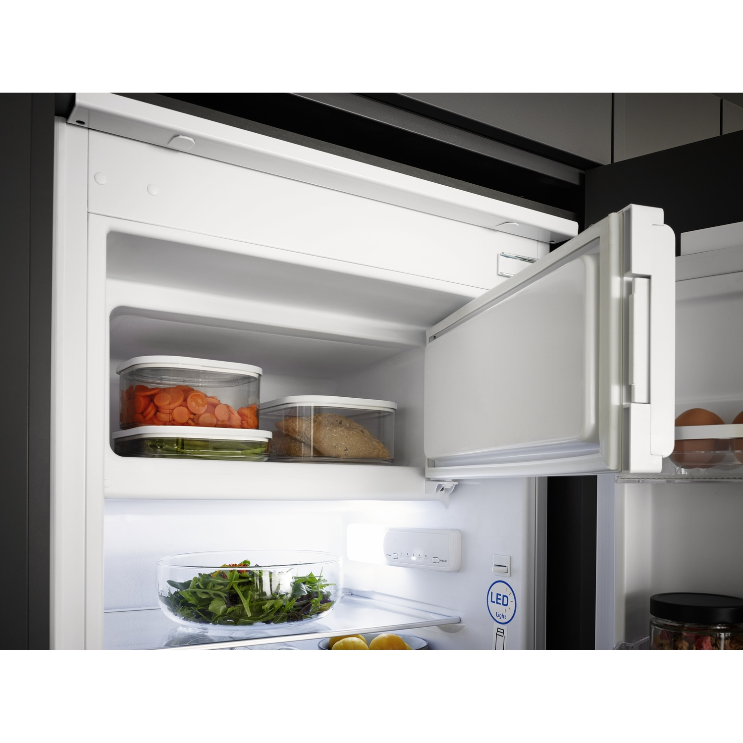 Réfrigérateur ouvert avec de la nourriture à l'intérieur. Carottes et pain dans des contenants, laitue dans un bol, œufs et un pot. Intérieur blanc.