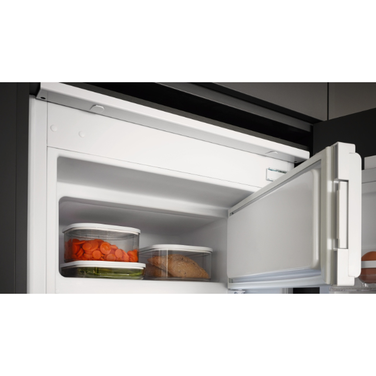 Réfrigérateur ouvert montrant des contenants transparents avec des carottes, du pain et des légumes verts. Intérieur et porte blancs. Fond sombre.