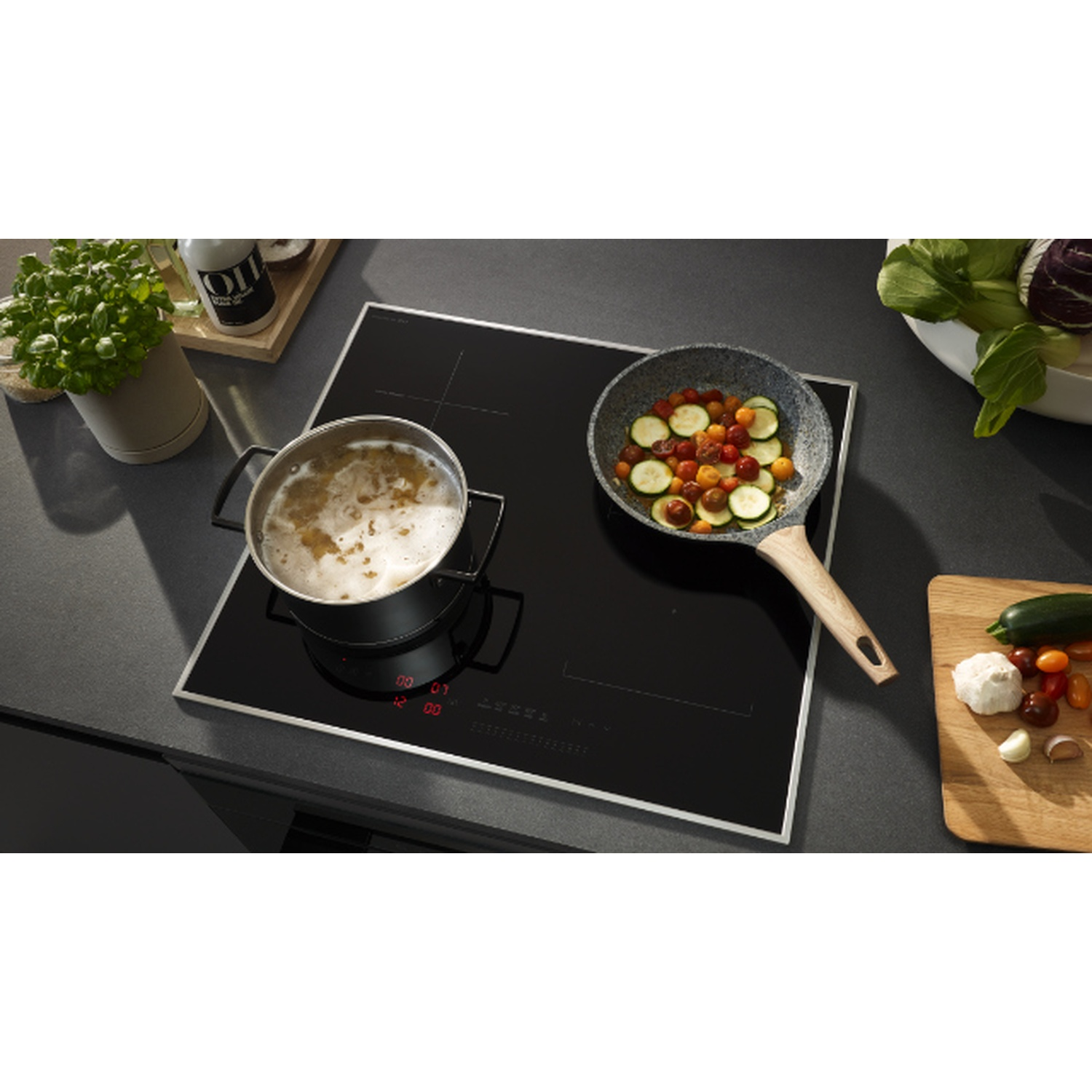 Une table de cuisson noire avec une casserole d'eau bouillante et une poêle de légumes. Une planche à découper avec de l'ail et une courgette à côté.