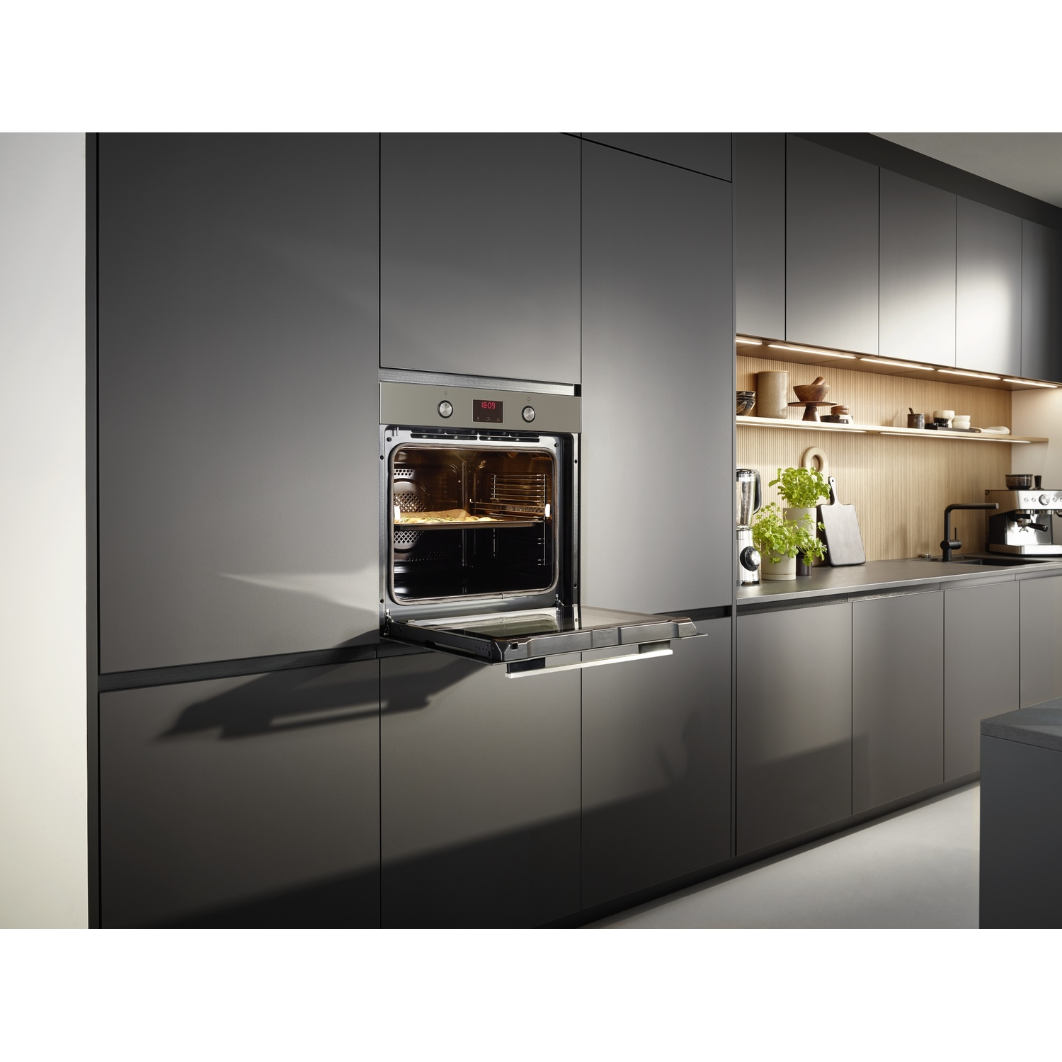 Four ouvert dans une cuisine moderne. À l'intérieur, des aliments cuisent. Des armoires gris foncé entourent le four. Arrière-plan : comptoir, plantes, cafetière.