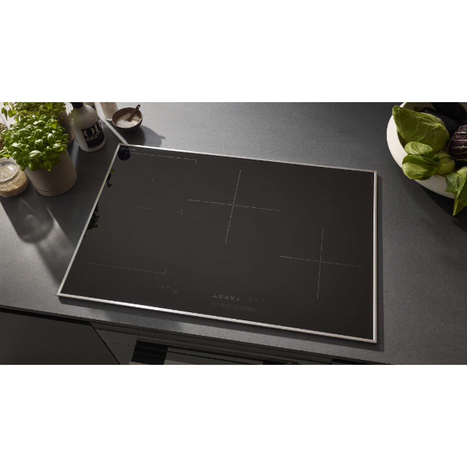Une table de cuisson à induction noire sur un comptoir sombre. Herbes, huile et un bol de légumes suggèrent une cuisine.