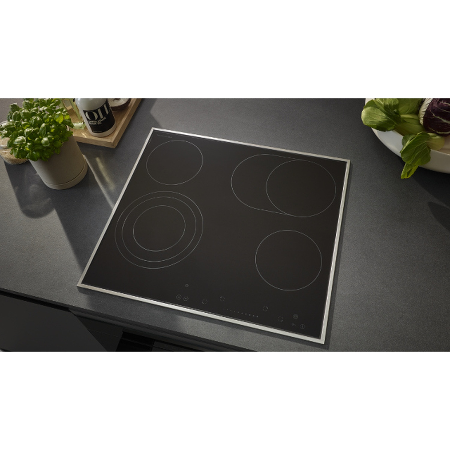 Une table de cuisson vitrocéramique noire avec quatre zones de cuisson circulaires. Herbes en pot, bouteilles d'huile et légumes en arrière-plan.