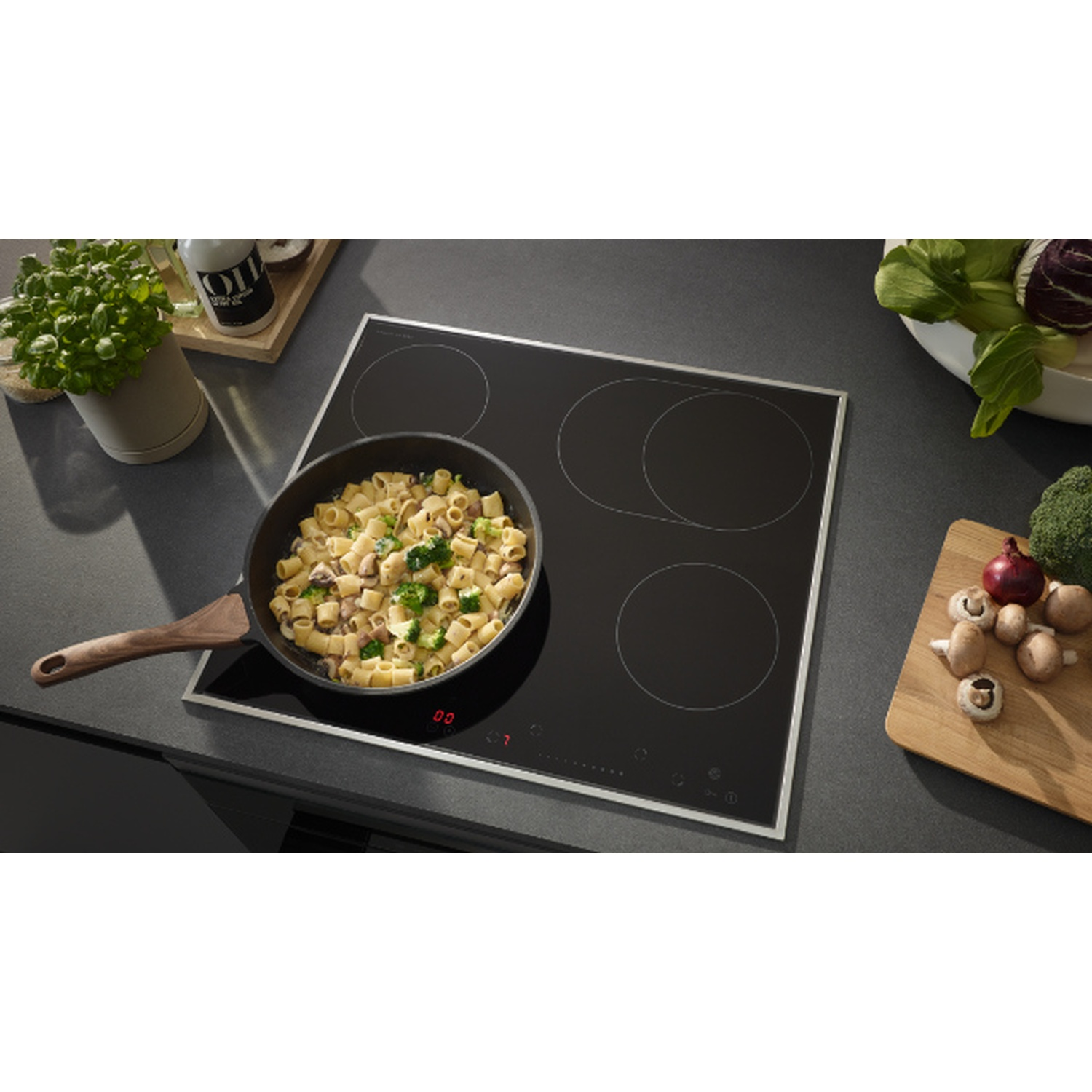 Une poêle de pâtes sur une table de cuisson noire, avec des légumes et une plante en arrière-plan, un cadre de cuisine moderne.