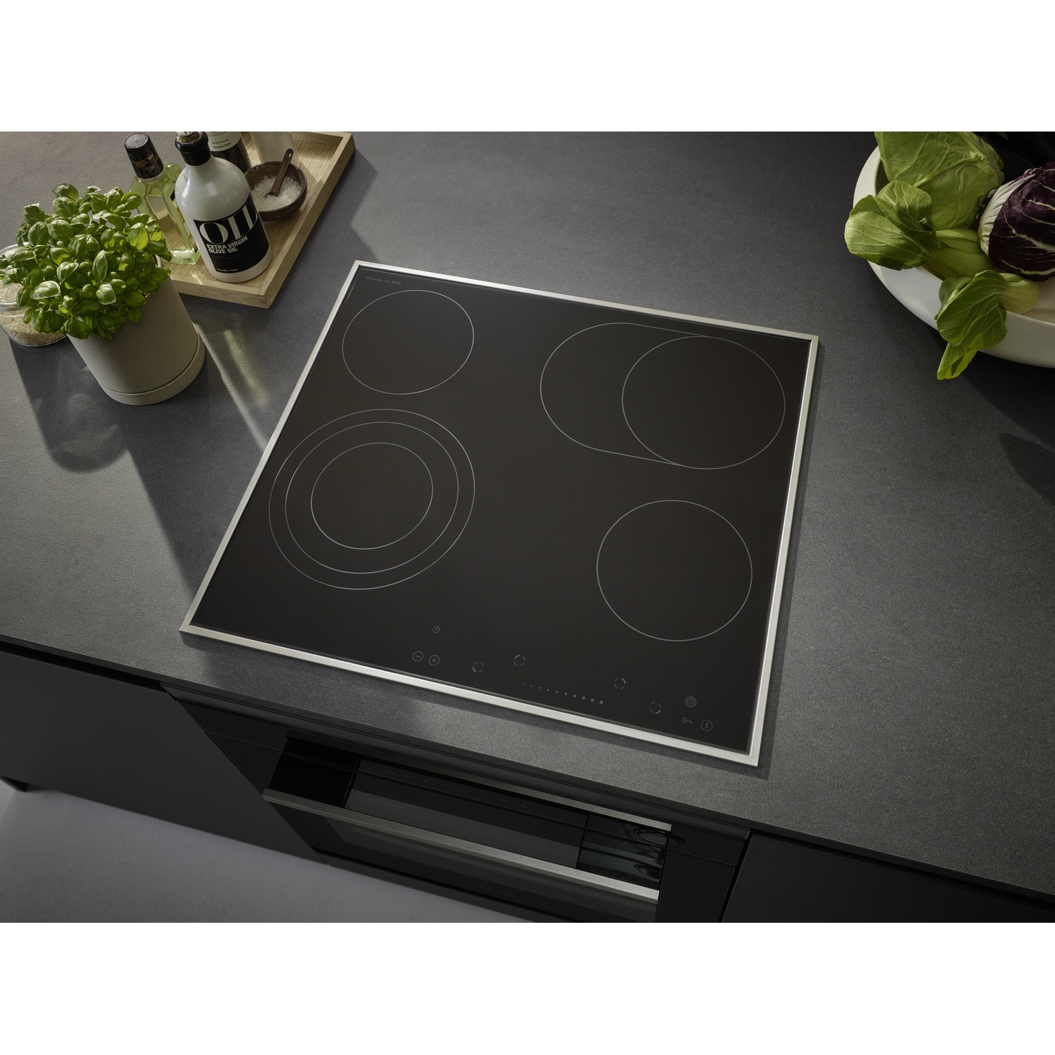 Une table de cuisson noire avec quatre zones de cuisson, entourée d'un cadre métallique. Légumes et bouteilles d'huile d'olive sur le comptoir.