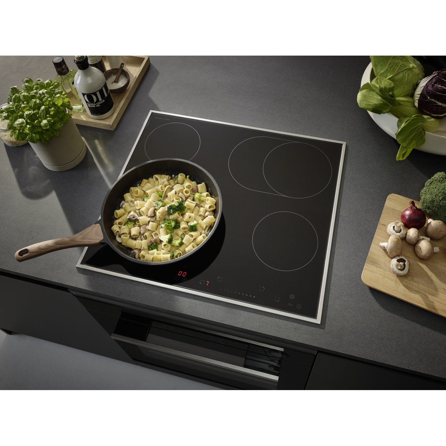 Scène de cuisine avec un plat de pâtes dans une poêle sur une plaque à induction noire. Légumes et bouteilles d'huile d'olive à proximité.