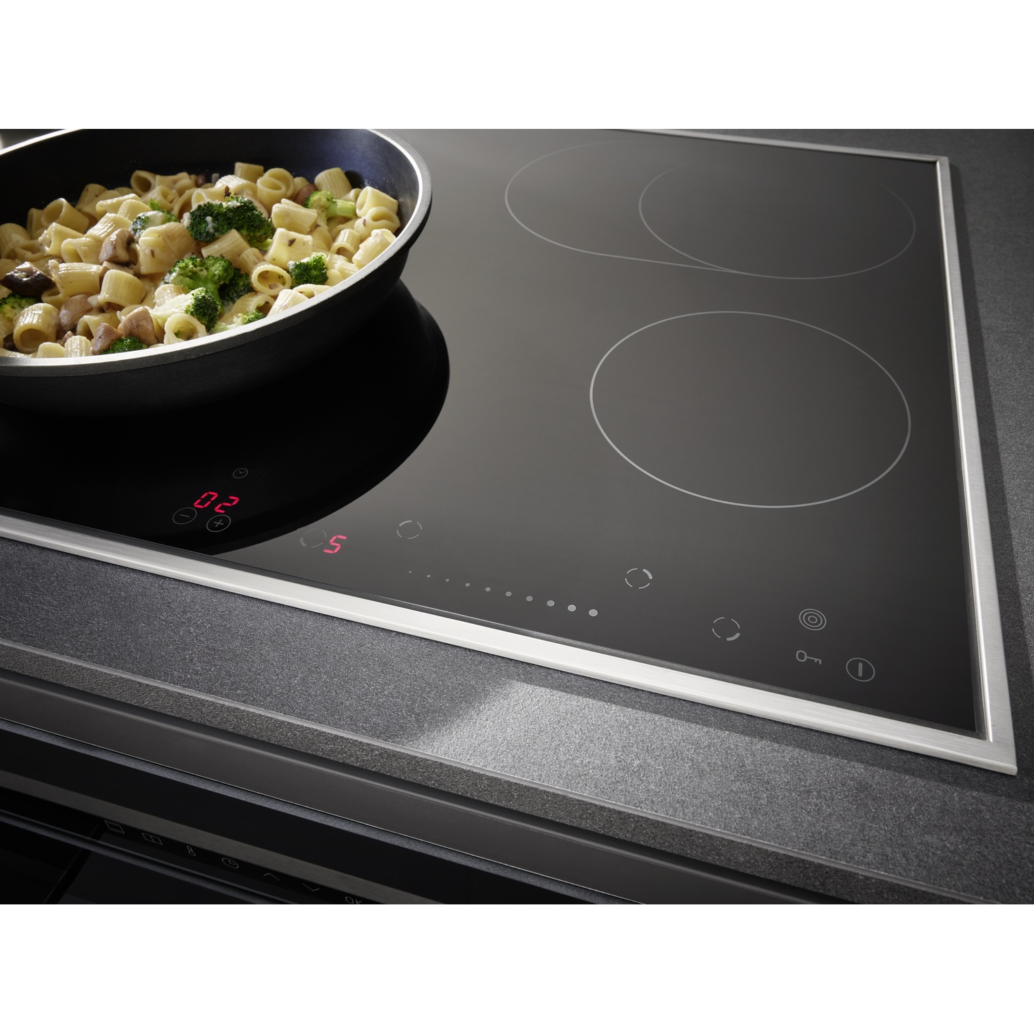 Une table de cuisson à induction noire avec une poêle de pâtes et de légumes. La table a trois zones de chauffe et un affichage numérique.