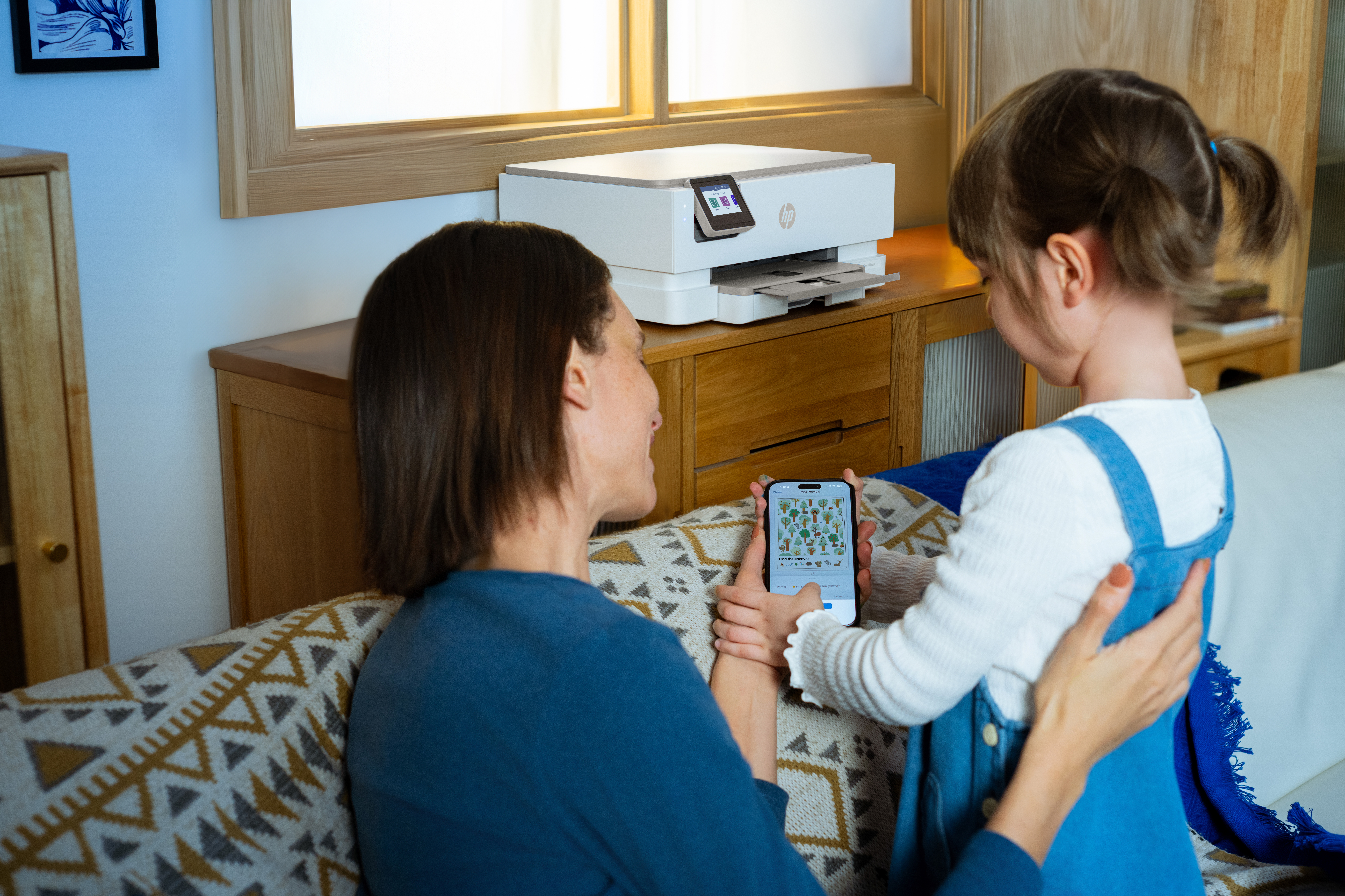 Kobieta i dziewczynka patrzą na telefon z aplikacją roślinną, obok drukarki na drewnianej szafce.