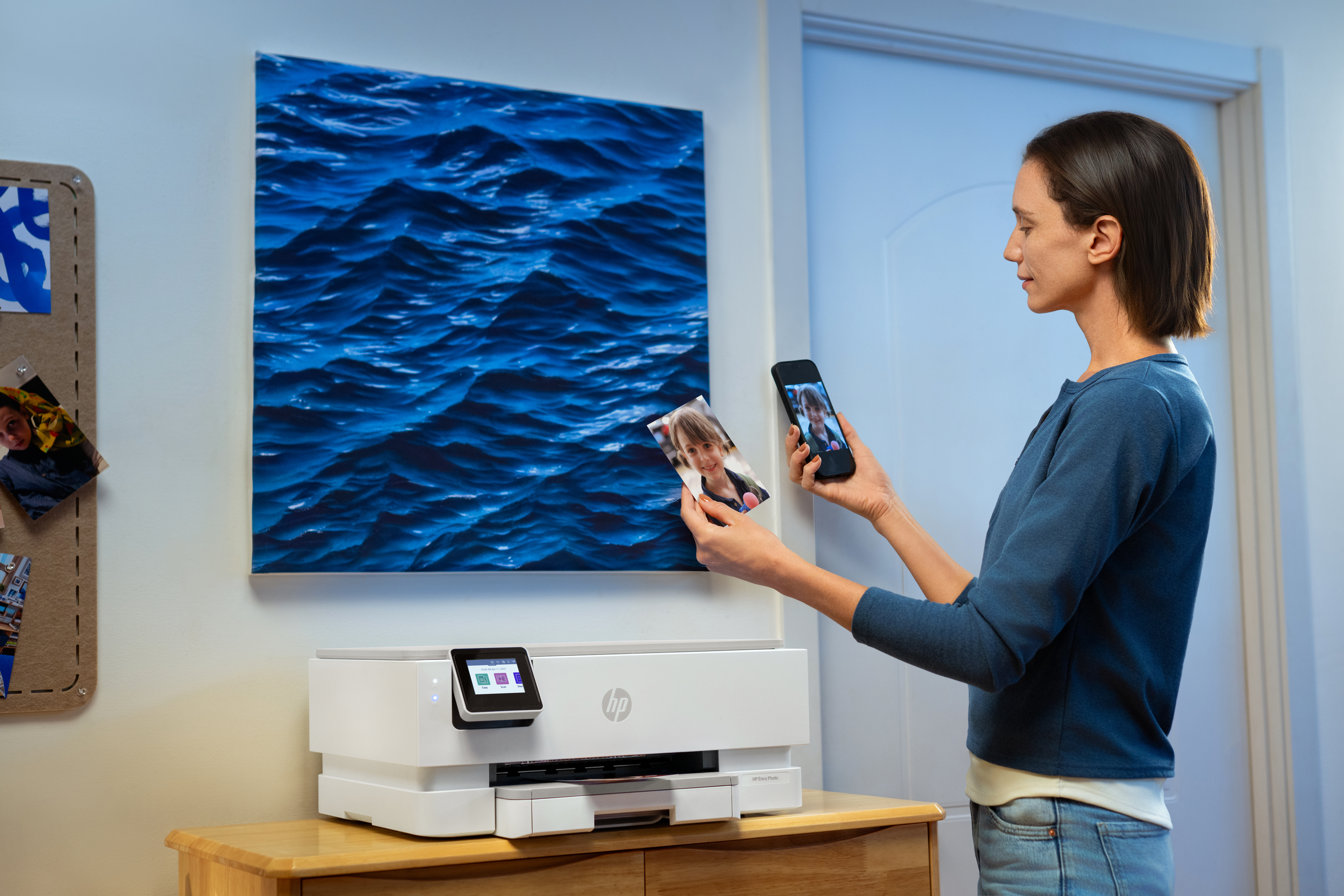 Kobieta trzyma zdjęcie i smartfon w pobliżu drukarki. Zdjęcie dziecka. Sztuka wody w kolorze niebieskim na ścianie.