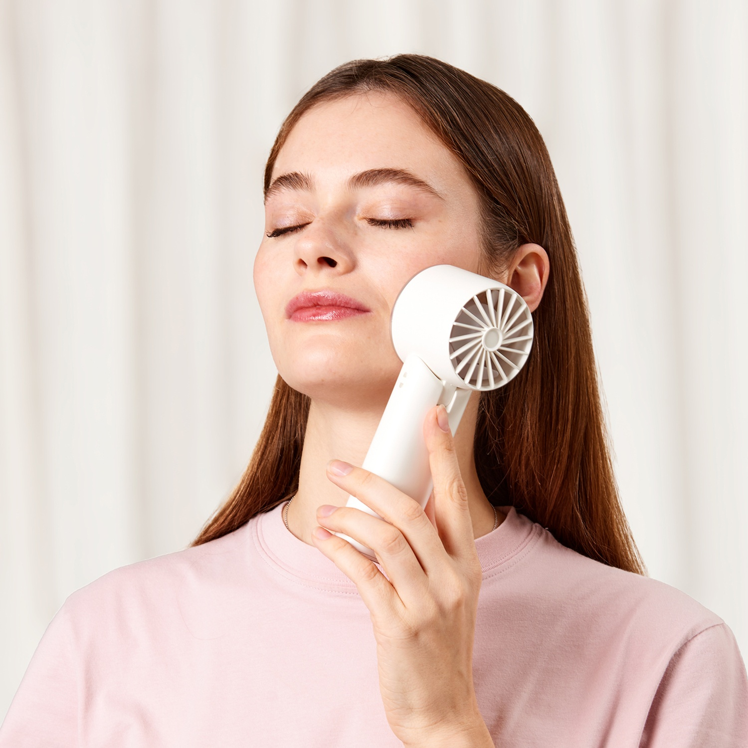Una donna con gli occhi chiusi tiene un ventilatore bianco sulla guancia. Indossa una maglietta rosa. Lo sfondo è una tenda bianca.