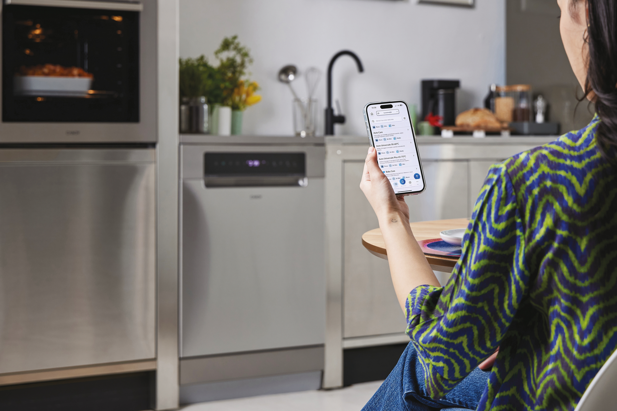 Una persona usa un teléfono en una cocina. El teléfono muestra opciones de lavavajillas. Se ven un horno y un lavavajillas.