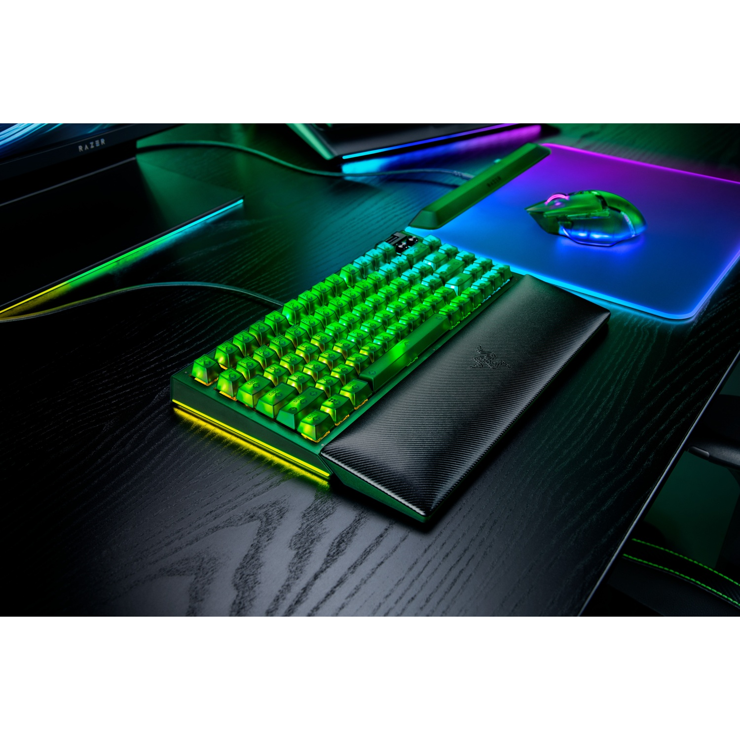 A gaming setup with a green-lit keyboard, mouse, and mousepad. The desk is black with a wood grain texture, with a Razer logo.