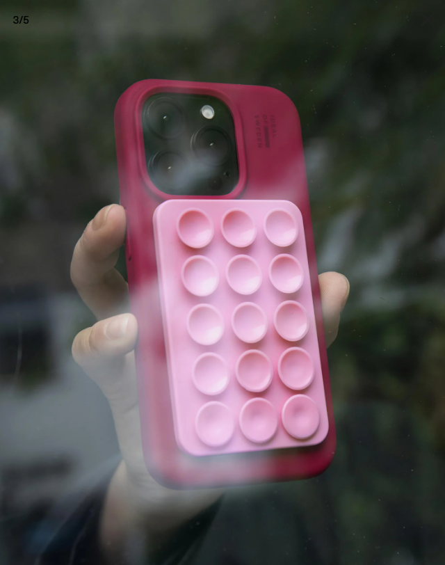 Una mano che tiene una custodia rosa per telefono con un pop-it attaccato. Lo sfondo è un fogliame verde sfocato. La custodia è tenuta in alto.