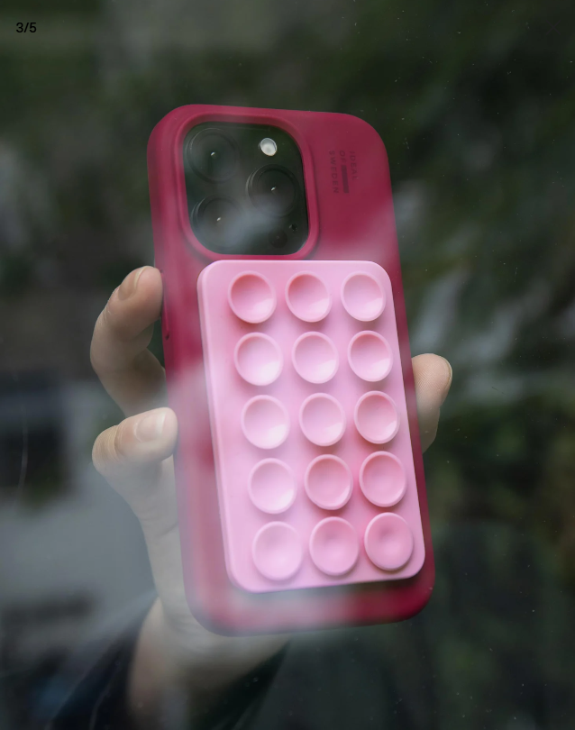 Una mano tiene una custodia rosa per telefono con un pop bubble attaccato. Lo sfondo è una vista sfocata di fogliame verde.