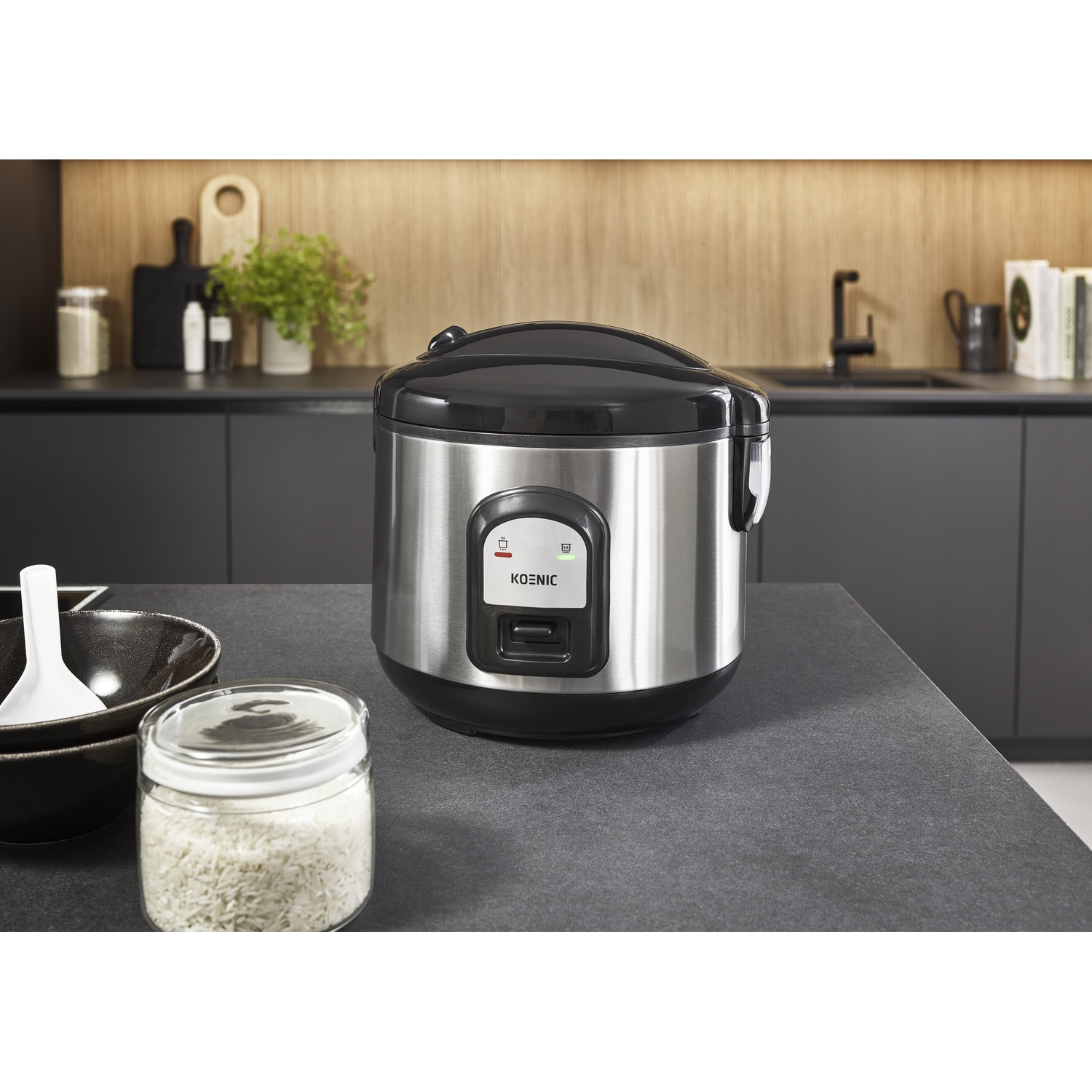 A stainless steel rice cooker sits on a dark countertop. A jar of rice, a bowl, and kitchen items are visible in the foreground.