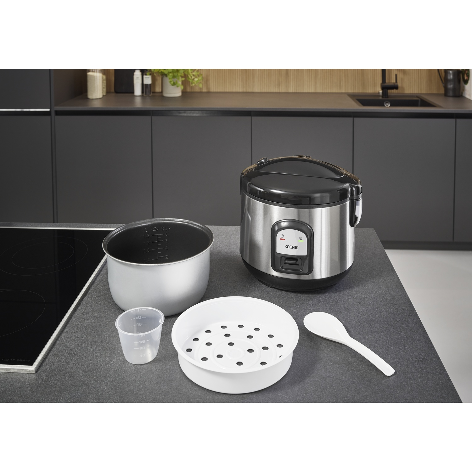 A rice cooker with accessories on a countertop. Includes a pot, measuring cup, steamer, spoon, and the cooker itself.