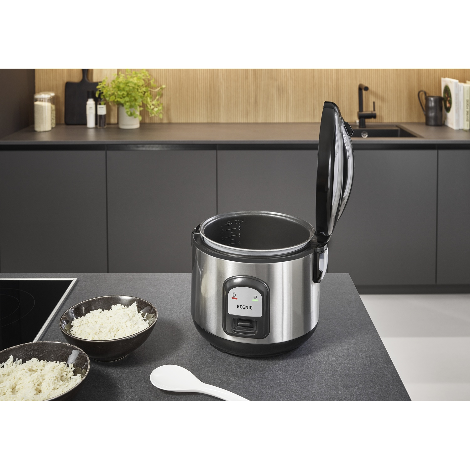 A rice cooker with an open lid, bowls of rice, and a spoon are on a counter. The background shows a kitchen.