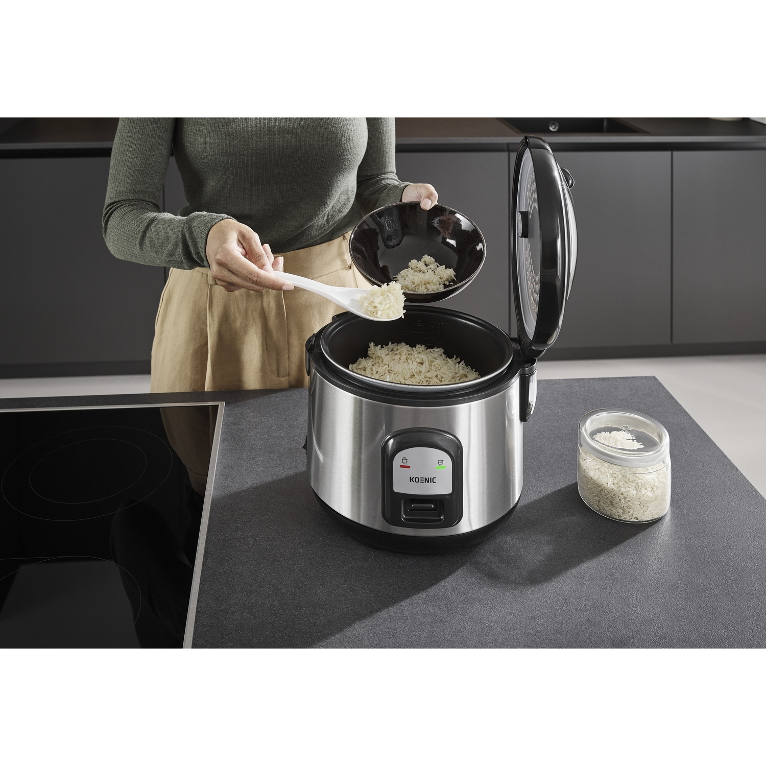 A person uses a spoon and bowl to pour rice into a rice cooker on a kitchen counter. A jar of rice is also present.