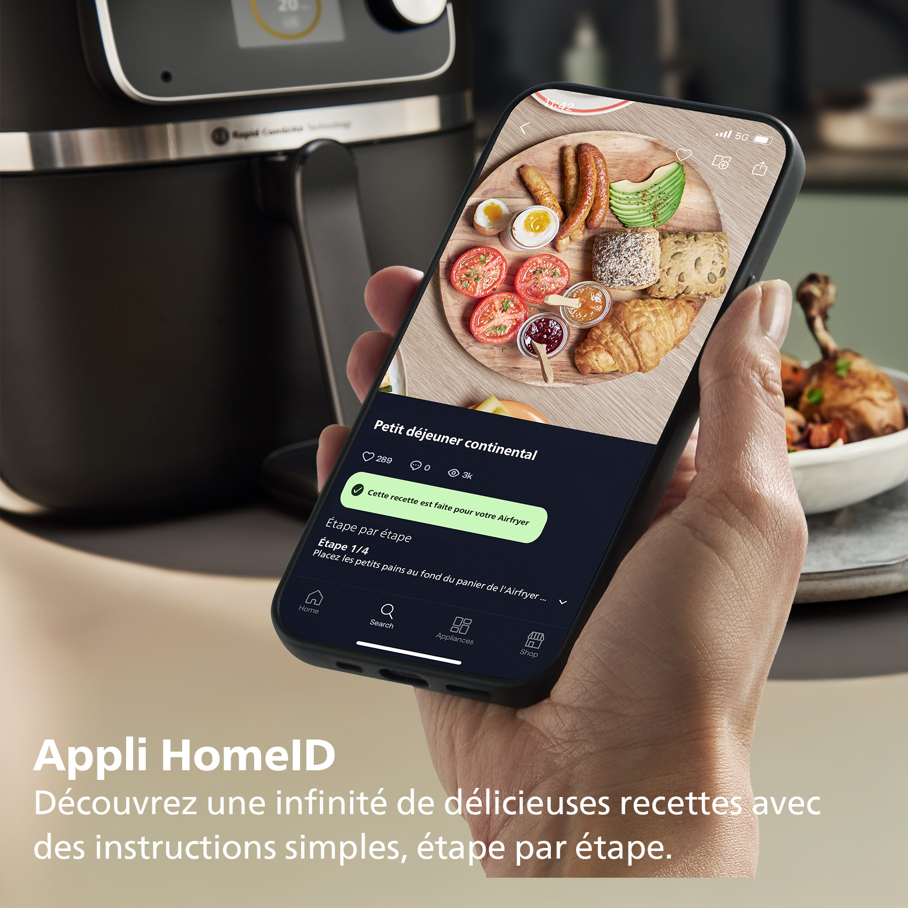 Une main tient un téléphone affichant une recette de petit-déjeuner continental. Un airfryer est en arrière-plan. Les aliments comprennent des œufs, du pain et des tomates.
