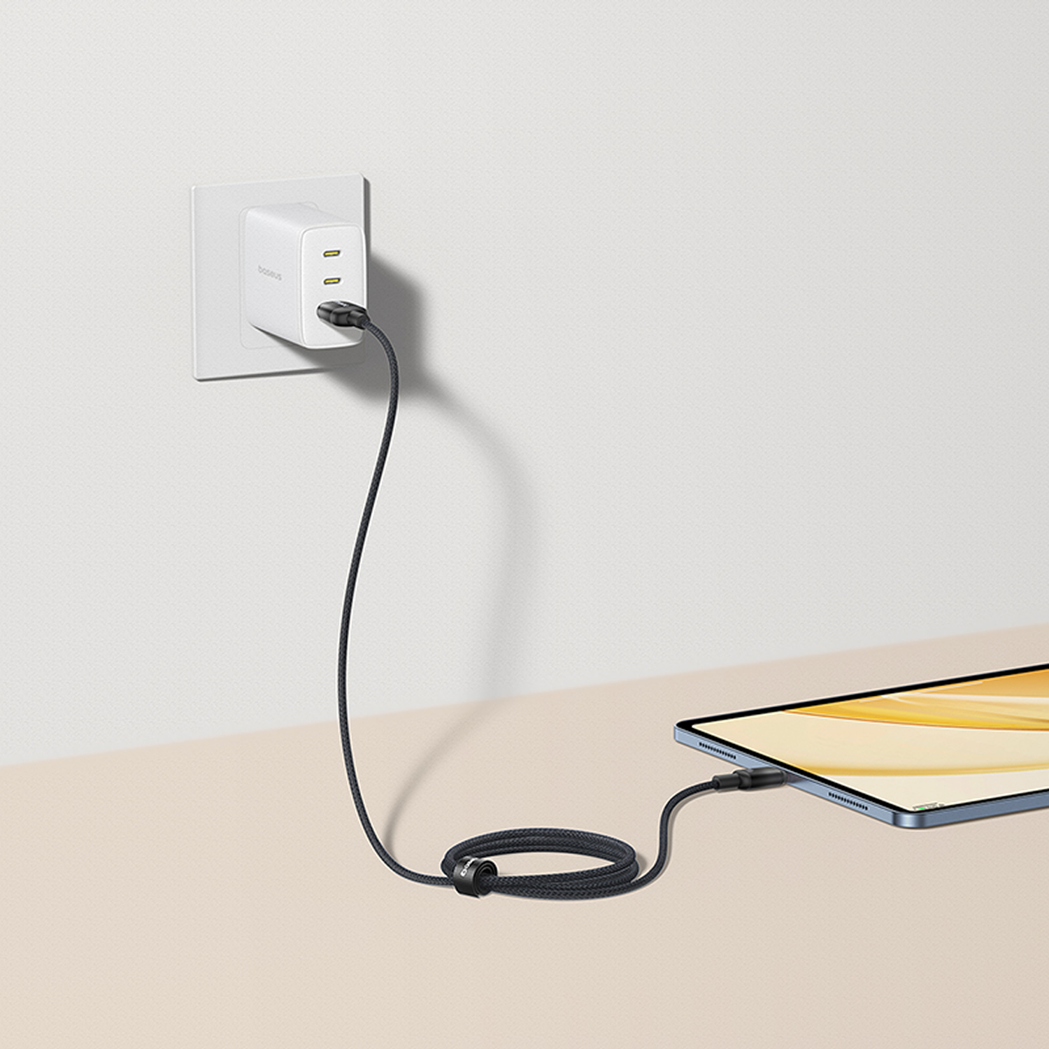 Un cargador blanco enchufado a la pared. Un cable negro conecta el cargador a una tableta sobre una superficie beige.