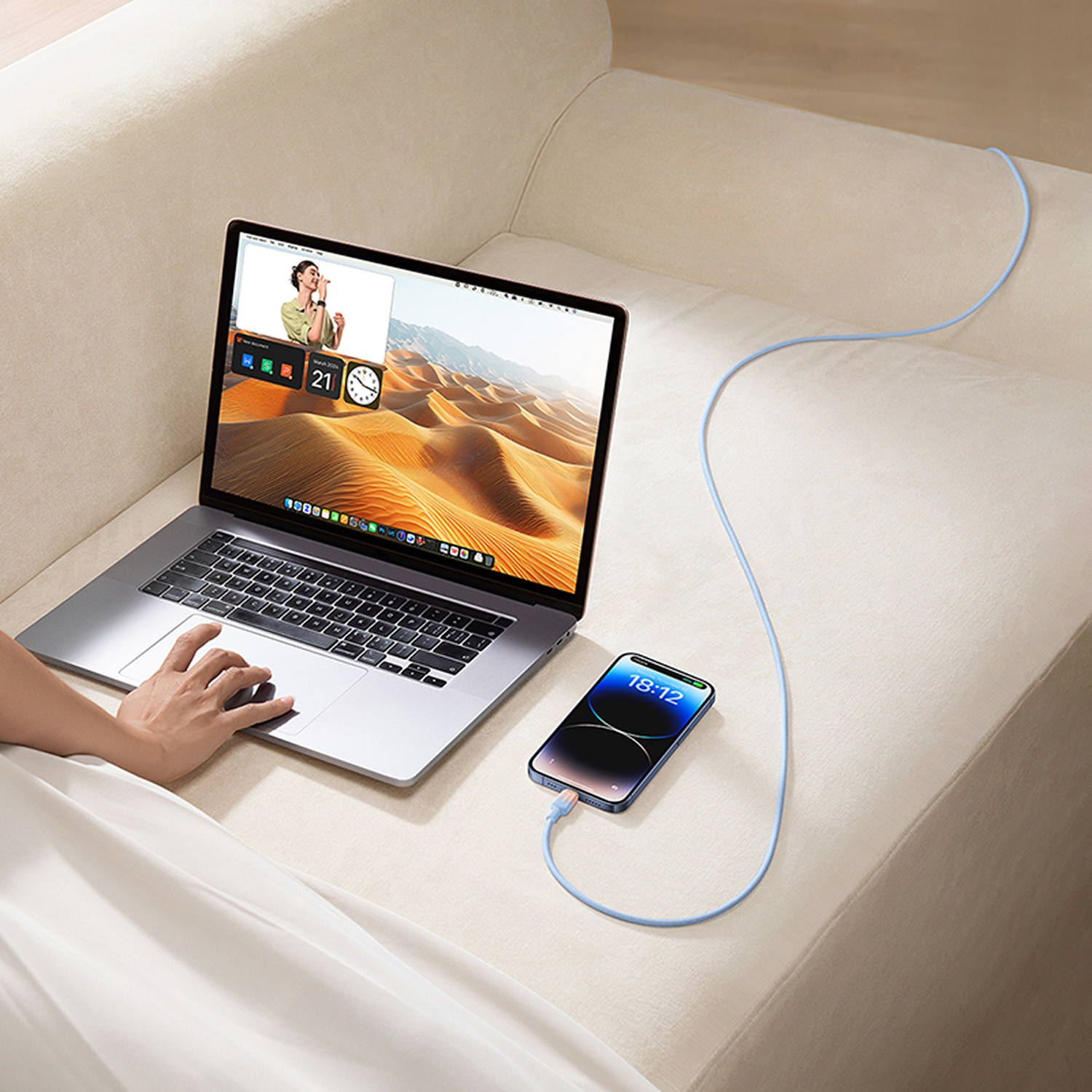 Ordenador portátil en un sofá beige con un teléfono cargando. El portátil muestra una videollamada y un fondo de pantalla desértico.