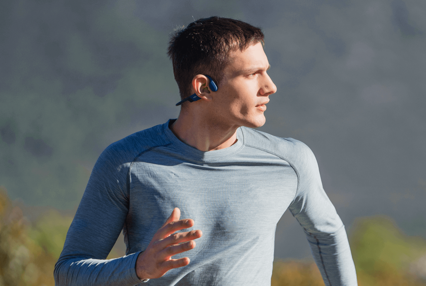 Mężczyzna biegnie na zewnątrz, ubrany w niebieską koszulę z długim rękawem i słuchawki. Tło to rozmyte góry.