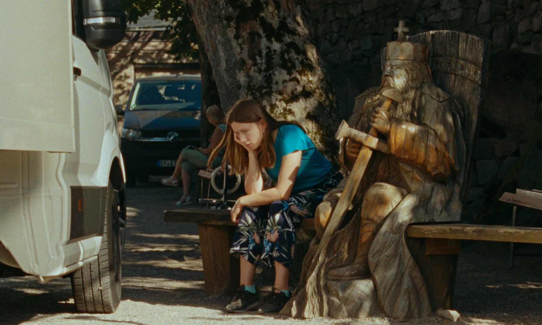 Eine Frau sitzt auf einer Bank neben einer Holzstatue. Links ist ein weißer Van geparkt. Ein Auto und Leute sind im Hintergrund.
