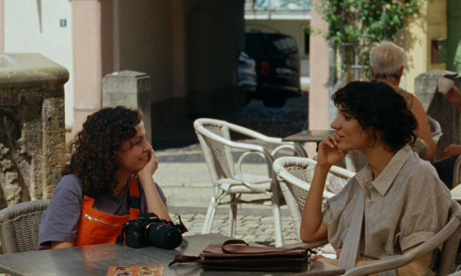Zwei Frauen an einem Cafétisch, lächelnd. Eine hat lockiges Haar, orangefarbene Latzhosen, Kamera. Die andere hat kurzes Haar, helles Hemd.