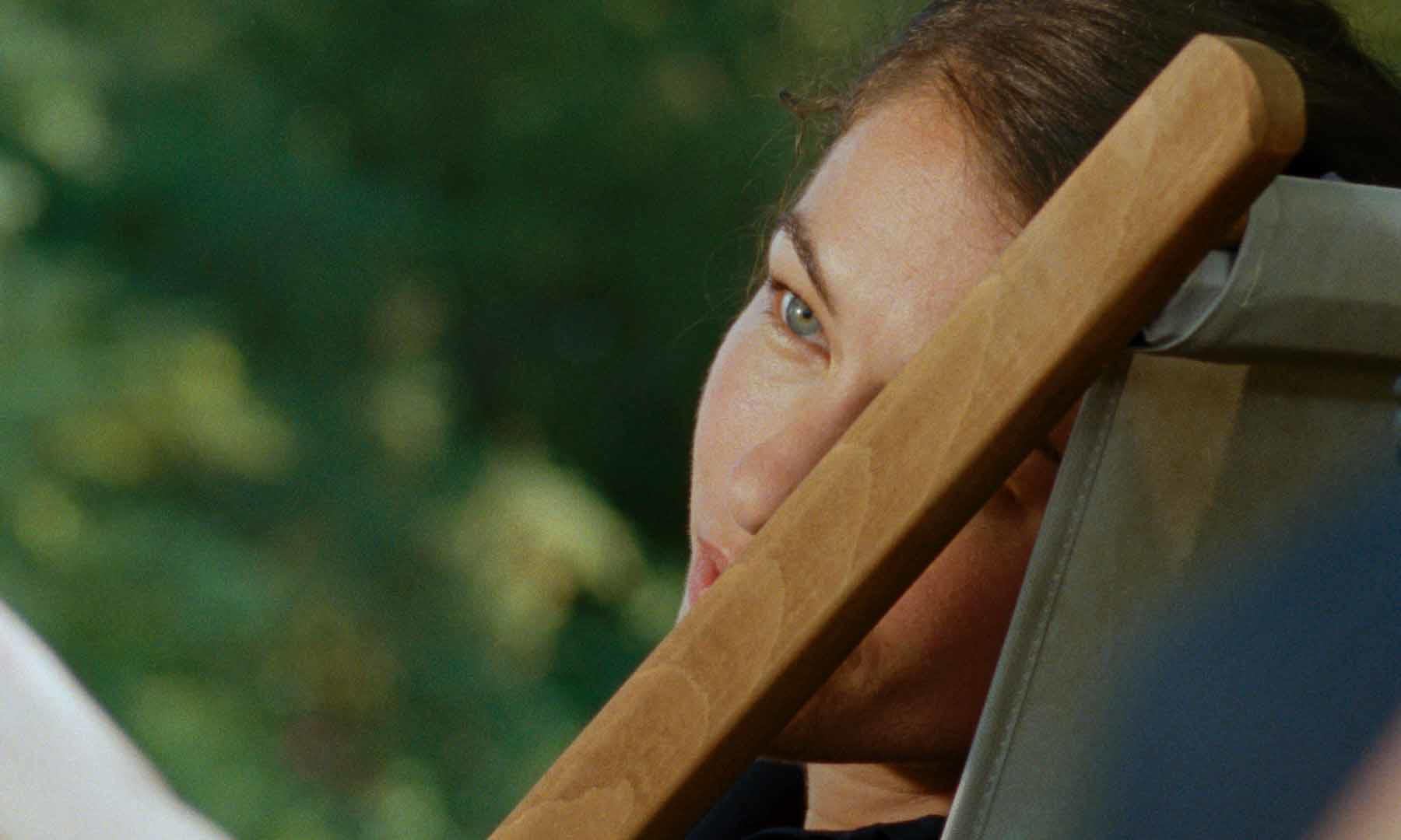 Eine Frau mit blauen Augen schaut zur Seite. Ein hölzerner Stuhlarmliegt davor. Grünes Laub im Hintergrund.