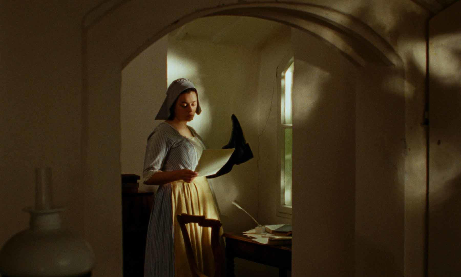Eine Frau in historischer Kleidung liest ein Papier in einem sonnendurchfluteten Raum. Sie steht in der Nähe eines Schreibtisches mit Papieren.